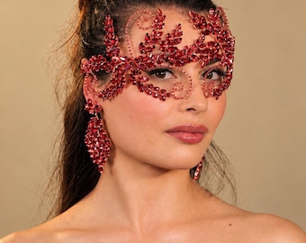 Puede incluir: Una máscara de cristal rojo con un intrincado patrón de hojas y enredaderas que cubre la parte superior del rostro y se extiende hasta las sienes. La máscara está adornada con numerosas piedras rojas facetadas e incluye pendientes a juego.