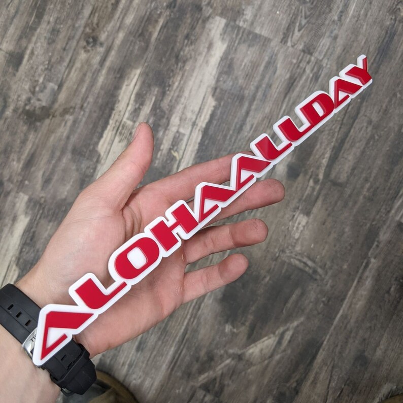 May include: A white plastic sticker with the word "ALOHA'LDAY" in red letters. The sticker is being held in a hand.