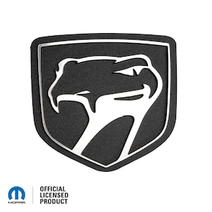 May include: Black and silver car badge featuring a snake head design. The badge is shaped like a shield and has the words "Official Licensed Product" printed below it.