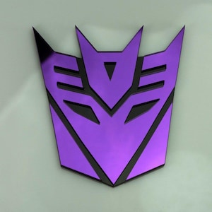 May include: Purple and black Transformers logo featuring a stylised robot face. The logo is a symbol of the Decepticons, a fictional group of robots from the Transformers franchise.