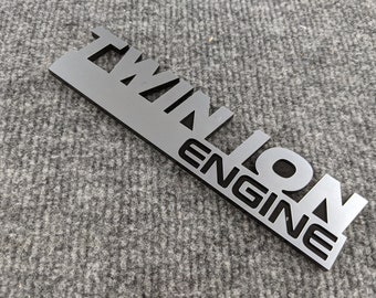 Może przedstawiać: Srebrny metalowy emblemat z napisem "Twin I/O" i "Engine" czarnymi literami.