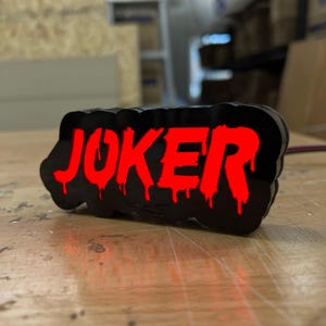 Puede incluir: Un letrero LED negro y rojo con la palabra "JOKER" en letras rojas que gotean. El letrero está iluminado y las letras están delineadas en negro.