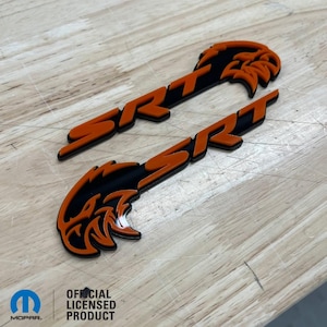 May include: Two orange and black SRT emblems with a stylized eagle head design. The emblems are made of a durable material and are perfect for adding a sporty touch to any vehicle.