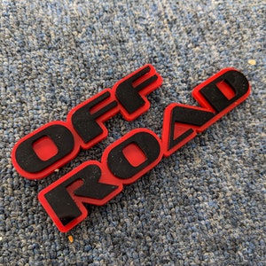 May include: Red plastic emblem with black lettering that reads "OFF ROAD".