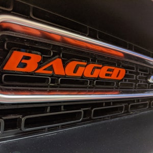 May include: A close-up of a black car grille with a silver trim. The word "BAGGED" is written in orange letters on the grille. The letters "GT" are also visible on the grille.