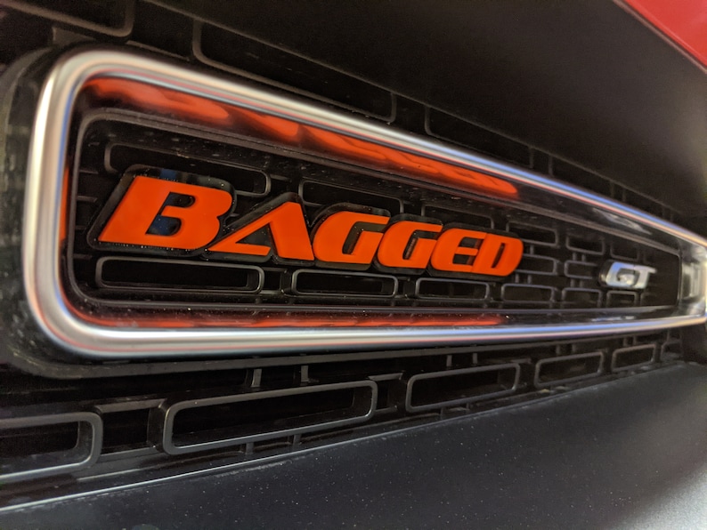 May include: A close-up of a black car grille with a silver trim. The word "BAGGED" is written in orange letters on the grille. The letters "GT" are also visible on the grille.