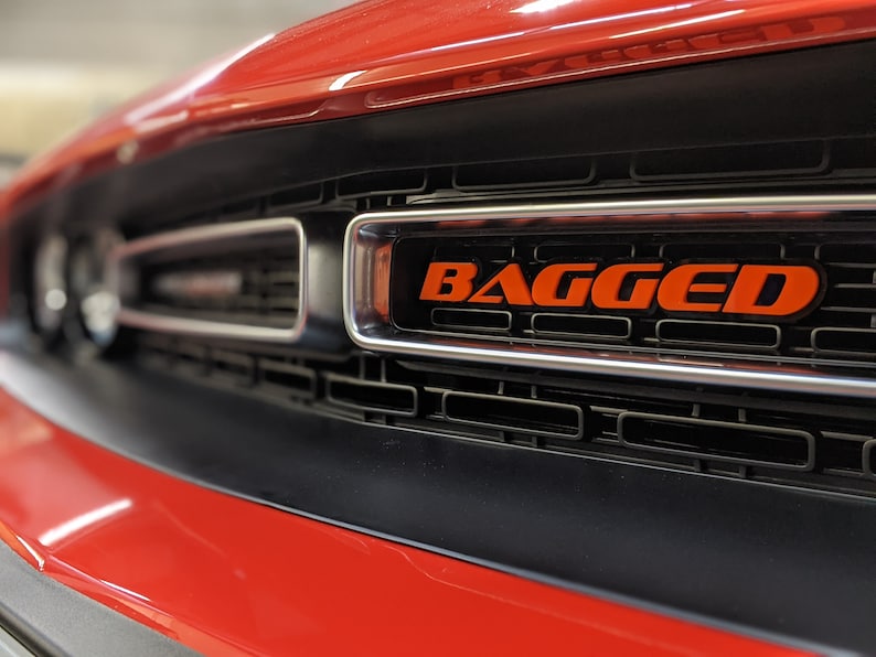May include: A close-up of a red car's front grille with a black mesh and a silver trim. The word "BAGGED" is written in orange letters on the grille.