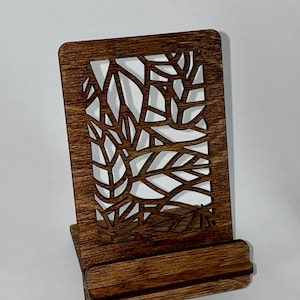 May include: A dark brown wooden phone stand with a decorative cutout design. The stand has a rectangular base and a raised back.