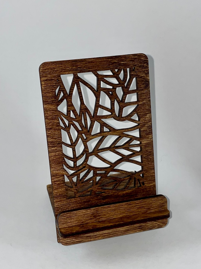 May include: A dark brown wooden phone stand with a decorative cutout design. The stand has a rectangular base and a raised back.