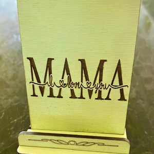 May include: A light green wooden sign with the word "MAMA" in black lettering and the phrase "I love you" with a heart symbol on either side of the word "love". The sign has a small stand on the bottom.