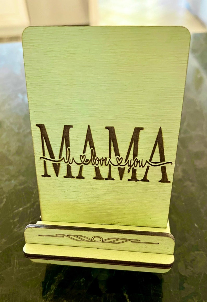May include: A light green wooden sign with the word "MAMA" in black lettering and the phrase "I love you" with a heart symbol on either side of the word "love". The sign has a small stand on the bottom.