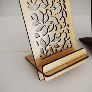 May include: A wooden phone stand with a decorative leaf pattern cut out. The stand is made of light brown wood and has a slot for holding a phone.