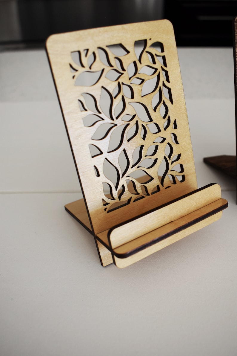 May include: A wooden phone stand with a decorative leaf pattern cut out. The stand is made of light brown wood and has a slot for holding a phone.