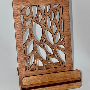 May include: A wooden phone stand with a cutout design of leaves. The stand is made of light brown wood and has a natural finish.