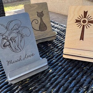 May include: Three wooden phone stands with different designs. The first stand has a black and white mushroom illustration and the text "Mush Love". The second stand has a black cat illustration. The third stand has a brown cross with a sunburst design.
