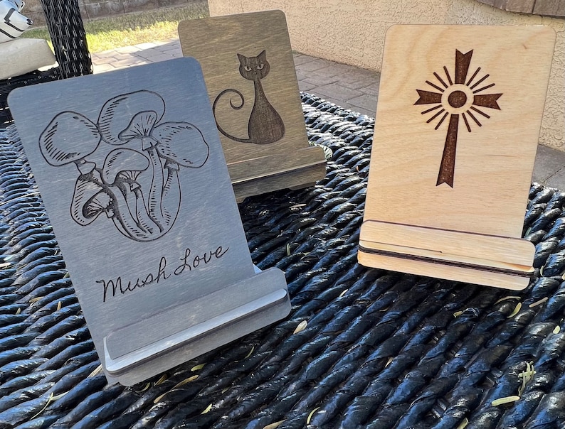 May include: Three wooden phone stands with different designs. The first stand has a black and white mushroom illustration and the text "Mush Love". The second stand has a black cat illustration. The third stand has a brown cross with a sunburst design.