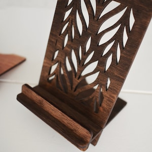 May include: A dark brown wooden phone stand with a decorative cutout design of leaves. The stand is angled to hold a phone upright.