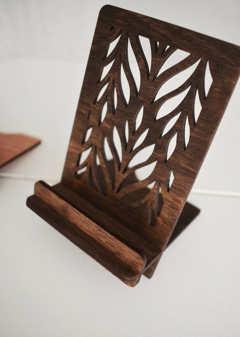May include: A dark brown wooden phone stand with a decorative cutout design of leaves. The stand is angled to hold a phone upright.