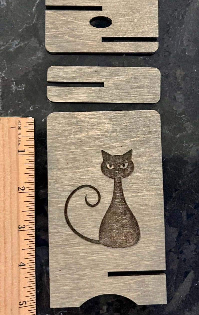 May include: A wooden coaster with a black cat design. The cat is sitting with its tail curled around its body. The coaster is made of wood with a light gray finish.
