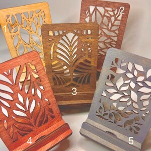 May include: Five wooden phone stands with different laser cut designs. The stands are stained in various colors, including natural wood, brown, red, and gray. The stands are numbered 1 through 5.