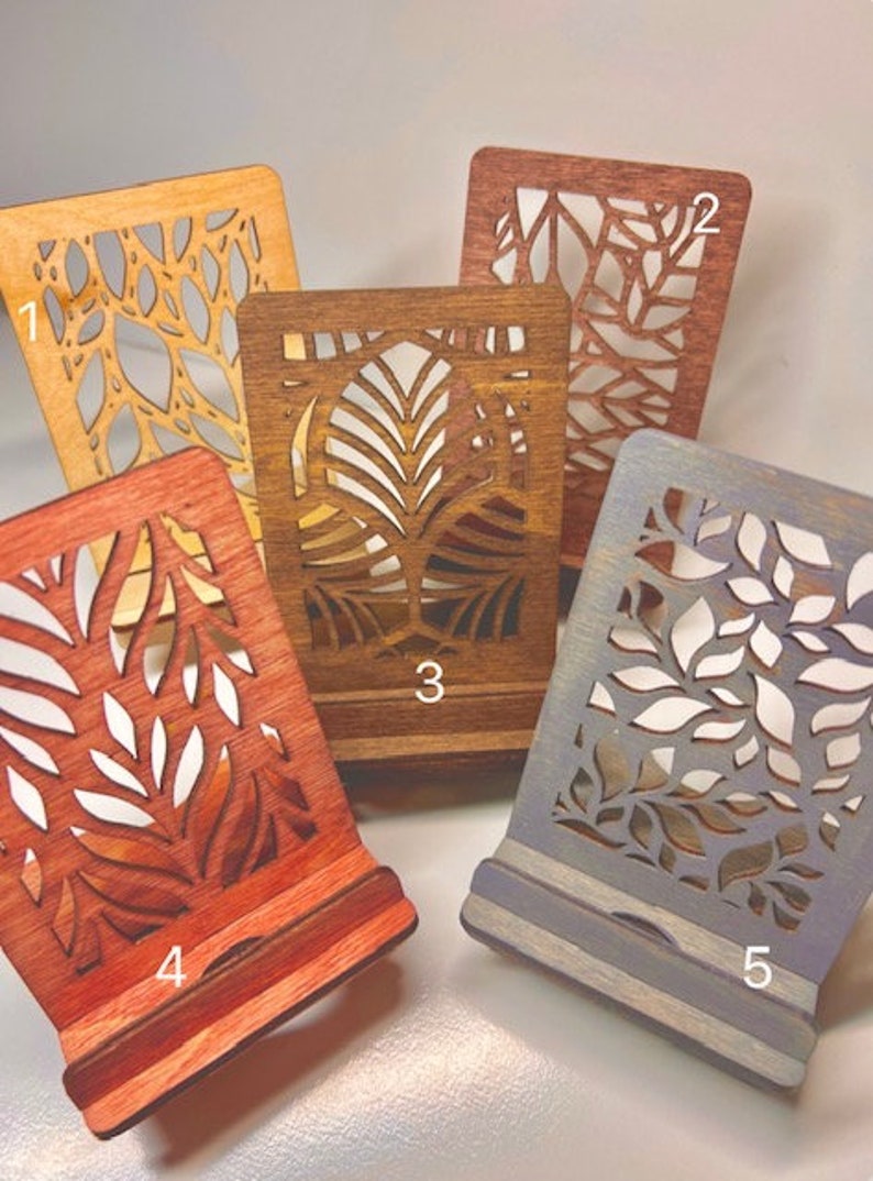 May include: Five wooden phone stands with different laser cut designs. The stands are stained in various colors, including natural wood, brown, red, and gray. The stands are numbered 1 through 5.