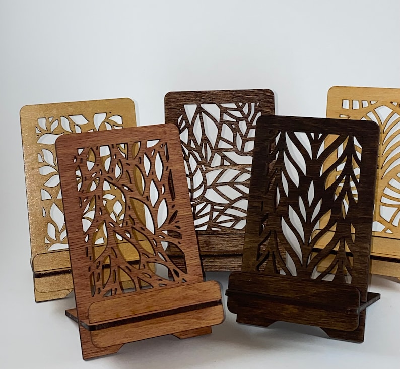 May include: Four wooden phone stands with intricate laser cut designs. The stands are made of different shades of brown wood and feature various leaf patterns.