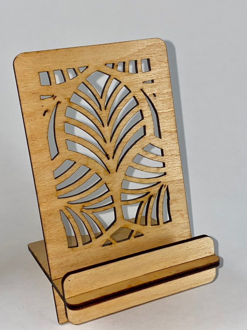 May include: A wooden phone stand with a decorative cutout design. The stand is made of light brown wood and has a simple, modern design.