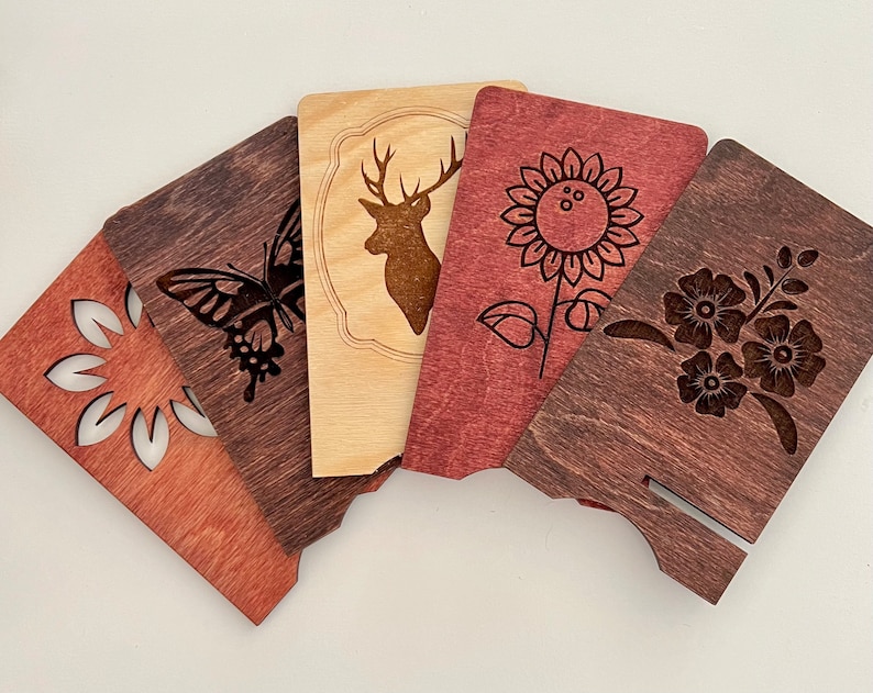 May include: Five wooden coasters with laser-cut designs. The coasters feature a butterfly, a deer, a sunflower, a floral design, and a simple geometric design. The coasters are stained in shades of brown and red.
