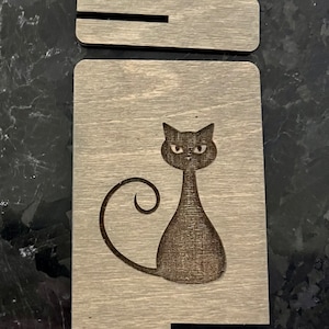 May include: A wooden sign with a black cat silhouette. The cat has a curled tail and is looking directly at the viewer. The sign is made of light brown wood with a wood grain pattern.