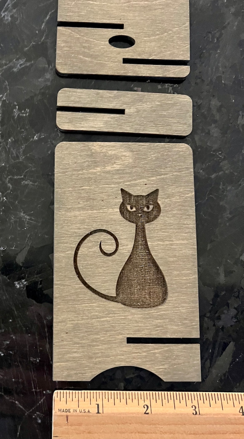 May include: A wooden sign with a black cat silhouette. The cat has a curled tail and is looking directly at the viewer. The sign is made of light brown wood with a wood grain pattern.