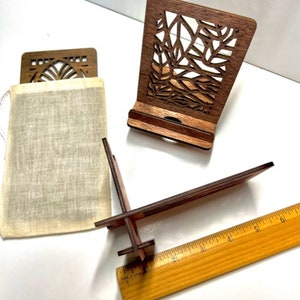 May include: Two wooden phone stands with intricate laser cut designs. One stand is in a natural wood color and the other is a darker brown. Both stands have a small pouch made of natural fabric.