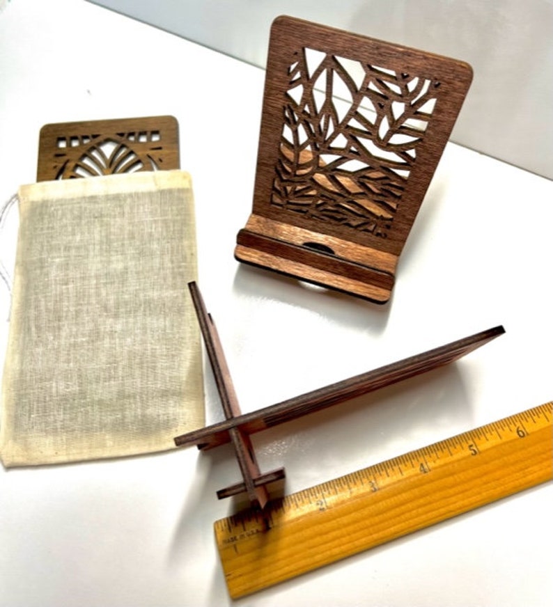 May include: Two wooden phone stands with intricate laser cut designs. One stand is in a natural wood color and the other is a darker brown. Both stands have a small pouch made of natural fabric.