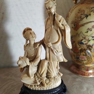 May include: A cream-colored figurine of two people, a man and a woman, standing on a black base. The man is wearing a long robe and has a beard. The woman is wearing a long dress and has her hair pulled back. The figurine is made of a material that looks like ivory.