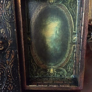 May include: An ornate wooden box with a hinged lid. The inside of the box is decorated with a green and gold oval frame with a faded image inside. The box is closed and the lid is visible.