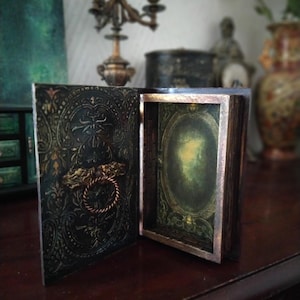 May include: A black and gold decorative box with an ornate design and a hinged lid. The box is open, revealing a green and gold interior with a mirror.