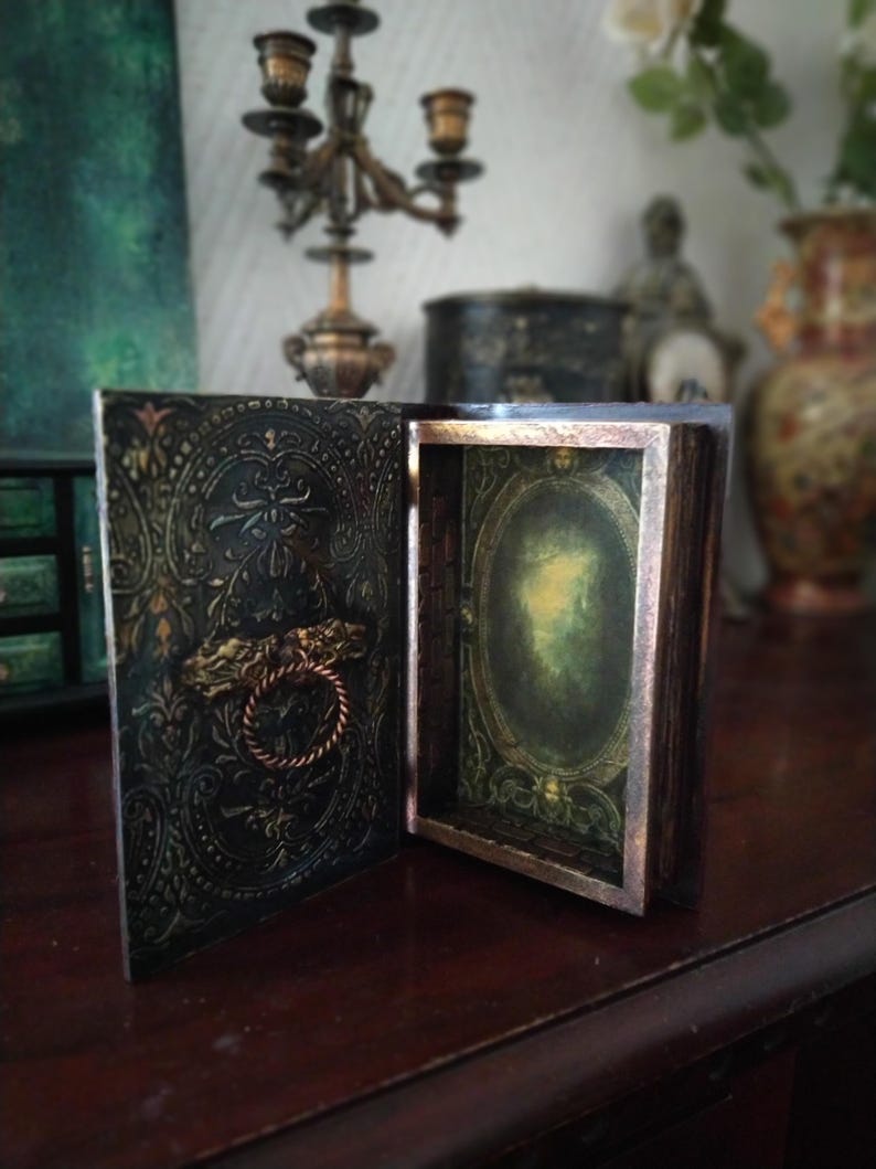 May include: A black and gold decorative box with an ornate design and a hinged lid. The box is open, revealing a green and gold interior with a mirror.
