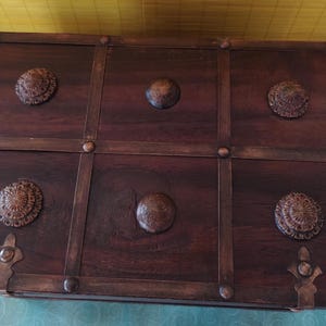 Op de afbeelding: Een donkerbruine houten kist met een decoratieve metalen rand en zes metalen accenten. De kist heeft een verweerde afwerking en is verdeeld in zes secties.
