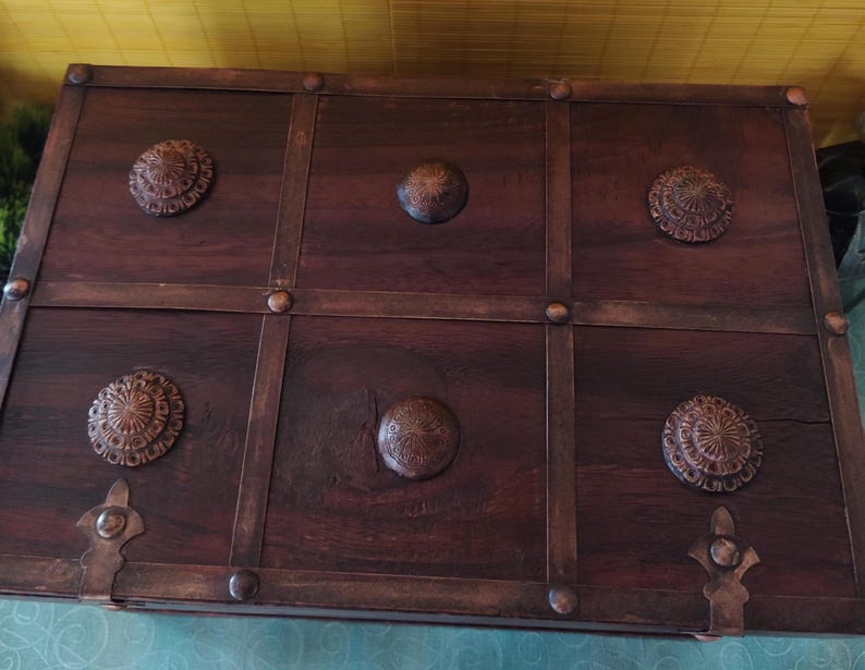 Op de afbeelding: Een donkerbruine houten kist met een decoratieve metalen rand en zes metalen accenten. De kist heeft een verweerde afwerking en is verdeeld in zes secties.