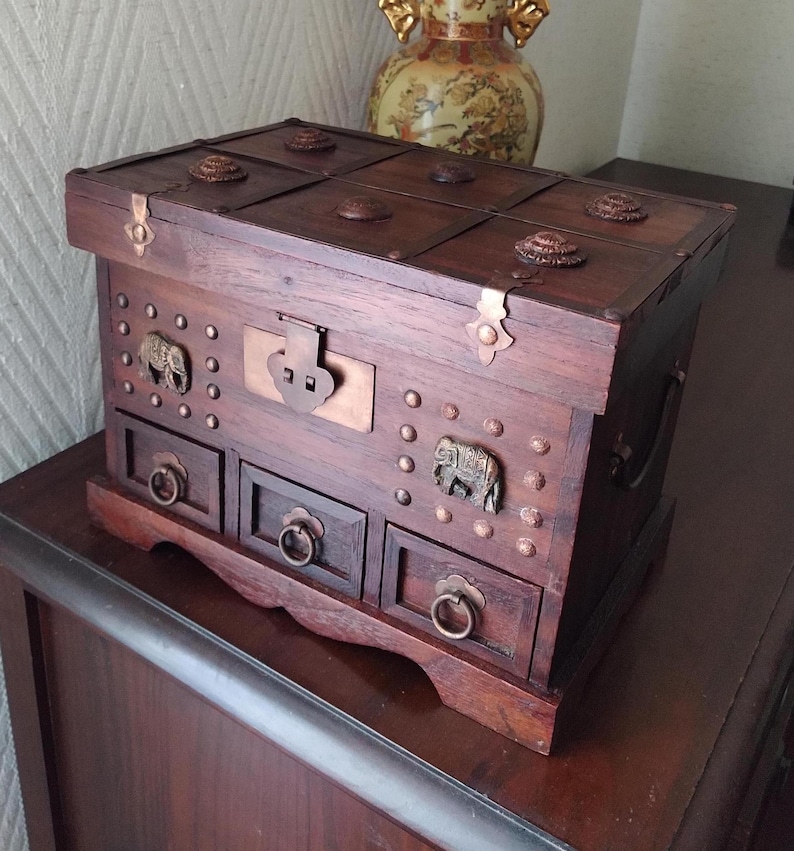 Op de afbeelding: Een houten juwelendoos met ingewikkelde details. De doos heeft een scharnierend deksel met een koperen sluiting en decoratieve messing accenten. De doos heeft vier laden met messing handvatten en is versierd met kleine messing studs en olifantenfiguren.