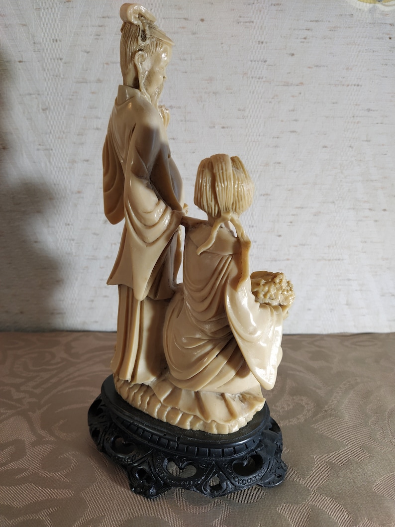 May include: A cream-colored ceramic figurine of a couple standing on a black base. The woman is holding a bouquet of flowers. The figurine is in a traditional Chinese style.