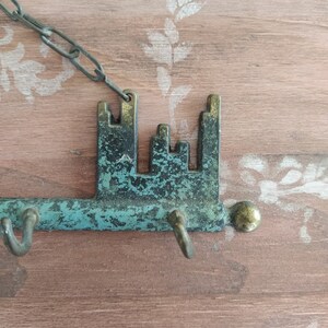 May include: A key-shaped metal key holder with a chain, featuring a distressed green and brown finish. The key holder has a decorative design of a city skyline and three hooks for hanging keys.