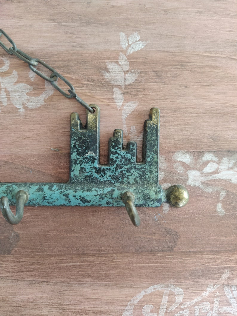 May include: A key-shaped metal key holder with a chain, featuring a distressed green and brown finish. The key holder has a decorative design of a city skyline and three hooks for hanging keys.
