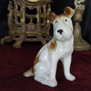 May include: A white porcelain figurine of a sitting dog with brown markings. The dog has a white body with brown ears, a brown spot on its back, and brown markings on its legs. The figurine is sitting on a red velvet surface.