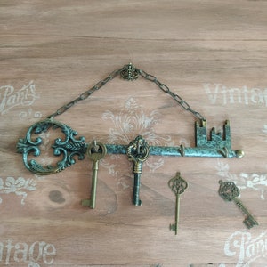 May include: A decorative key-shaped wall hook with a chain and a small crown at the top. The hook is painted in a teal green color and has a rustic, vintage look. Four antique-style keys are hanging from the hook.