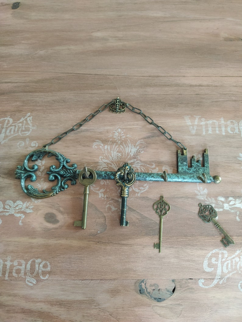 May include: A decorative key-shaped wall hook with a chain and a small crown at the top. The hook is painted in a teal green color and has a rustic, vintage look. Four antique-style keys are hanging from the hook.