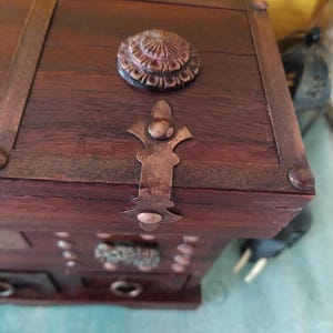 Op de afbeelding: Een houten doos met een scharnierend deksel en decoratieve metalen accenten. De doos heeft een verweerde bruine afwerking en beschikt over een metalen sluiting met een fleur-de-lis-ontwerp.