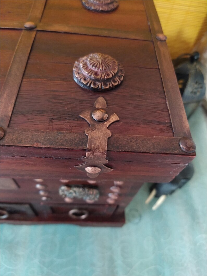 Op de afbeelding: Een houten doos met een scharnierend deksel en decoratieve metalen accenten. De doos heeft een verweerde bruine afwerking en beschikt over een metalen sluiting met een fleur-de-lis-ontwerp.