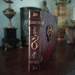 May include: A burgundy leather-bound journal with gold embossed details. The cover features a dragon design and the spine has intricate floral patterns.