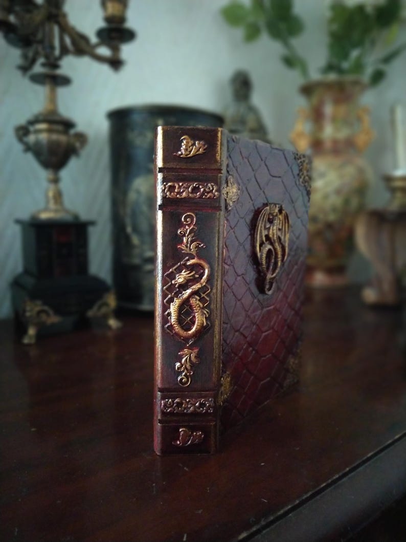 May include: A burgundy leather-bound journal with gold embossed details. The cover features a dragon design and the spine has intricate floral patterns.