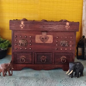 Op de afbeelding: Een donkerbruine houten kist met drie laden en decoratieve olifantenaccenten. De kist heeft een scharnierend deksel met een metalen sluiting en koperen accenten. De kist staat op een blauw-groen gepatroneerd oppervlak.